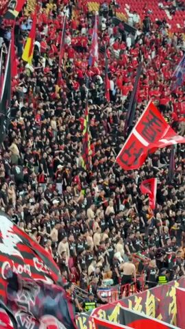 A large crowd fills the stands of a stadium, a mix of red and black clothing dominating the view. Many individuals are waving flags, some red with white characters, others black and red with various designs. A few people are shirtless. The camera moves, showing more of the stadium's seating, which is mostly red, and then pans down to a green field. There's a sense of a lively, possibly sporting, event in progress.