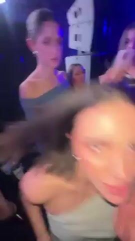 The video, shot from a first-person perspective, shows a woman dancing in a crowded environment, possibly a club or party, under blue lighting. She is wearing an off-the-shoulder top. Other people are visible in the background, moving and dancing as well. The camera is shaky and often blurs due to the woman's movements. The music playing appears to be upbeat and dance-oriented. The focus is primarily on the woman and her immediate surroundings.