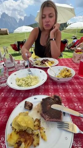 A young woman with blonde hair is sitting at a red and white checkered table outdoors. She is wearing a black tank top and yellow pants. She is eating a meal from a white plate with a fork and knife. In front of her are several plates of food, including steak, potatoes, and salad. There are also drinks on the table, including a glass of water and a red beverage. In the background, there are mountains, trees, and other people sitting in chairs. The woman appears to be in a restaurant or cafe.
