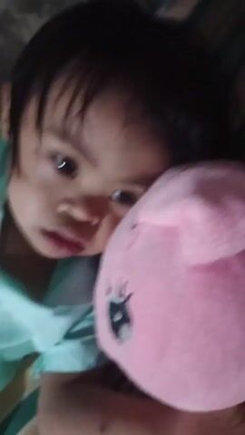 A young child is lying down, cradling a pink plush toy with a cartoon eye on it. The child is wearing a light blue shirt and has short dark hair. The child is looking upwards. The background appears to be a striped surface.