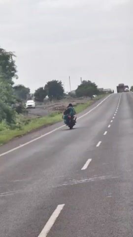 A motorcycle carrying two people is traveling on a paved road. The person in the front is driving the motorcycle. The person in the back is wearing a blue garment. The road is lined with grass and trees. In the distance, there are buildings and a power line. The sky is overcast.