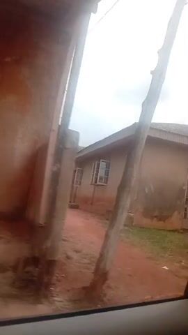 The video captures a moving shot of a modest building, likely a house or small commercial structure, with a corrugated metal roof. The exterior walls appear to be unpainted concrete or stucco, showing signs of age and weathering. There are several windows visible, some with metal bars for security. The front of the building features large, arched openings covered with similar barred gates. A sign is affixed to one of these gates, though the text is partially obscured and difficult to read completely, it appears to be an advertisement for a property, possibly a "2 BEDROOM FLAT." The surrounding environment is characterized by a dirt or gravel road, patchy grass, and overgrown vegetation. In the background, a few water tanks are visible on top of another structure, suggesting a residential or utilitarian area. The sky is overcast with clouds. The camera perspective suggests the video is being recorded from inside a vehicle, looking out through a window, as indicated by the partial view of a car door and interior trim. There are no people clearly visible or engaged in any specific actions within the frame. The overall impression is one of a developing or somewhat neglected urban or semi-urban setting.