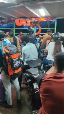 The video shows a crowded ferry. People are sitting on motorcycles and standing. Some people are wearing life jackets. The ferry appears to be moving on water at dusk. There is a fire extinguisher visible on the ferry.