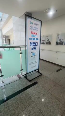 The video opens with a close-up of a vertical sign standing on the floor. The sign is white with blue and red text, and an image of a computer screen. The text appears to be in Korean and mentions "Happy Start" and "Computer Education." The camera then pans up and to the left, revealing a glass balcony with a metal railing. Beyond the balcony, a multi-story atrium is visible with a glass ceiling. Several flags of different countries, including the Union Jack, are displayed on poles throughout the atrium's ground floor. The view looks down into the atrium, showing an open space with tiled flooring and various doors and windows along the walls. Buildings and trees can be seen outside through the glass ceiling and walls. The camera continues to pan, offering a wider view of the atrium and its surroundings. There are no people visible in the immediate foreground, but a person in dark clothing can be seen briefly near a doorway in the distance. The overall environment is an indoor public space, likely a lobby or common area, with an emphasis on natural light and an international theme indicated by the flags.