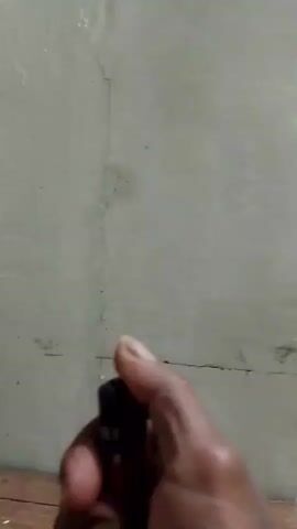 A close-up shot shows a hand holding a black lighter. The lighter is ignited, producing a small flame with blue and yellow colors. The background is a textured, light-colored wall with some darker marks and lines. The flame flickers for a moment before being extinguished, and the lighter is released. The shot remains focused on the hand and the lighter.