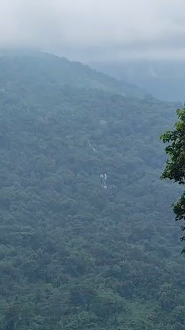A wide shot shows a lush green mountain covered in dense trees. A small waterfall is visible in the distance, cascading down the mountainside. The sky is overcast and hazy, with clouds obscuring the tops of the mountains. The scene is tranquil and serene, with the sounds of nature all around.