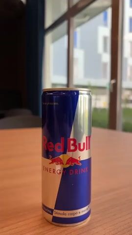 A can of Red Bull energy drink sits on a light-brown wooden table. The can's design is the classic blue and silver with the red bulls logo. The text on the can appears to be partially in Italian, with "Stimola corpo e mente" visible at the bottom.  Behind the can, out of focus, is a window showing a blurred view of buildings and greenery. There is also a hint of a dark blue curtain and a dark piece of furniture in the background. The camera angle is slightly above the can, looking down at it. The lighting is bright and natural.