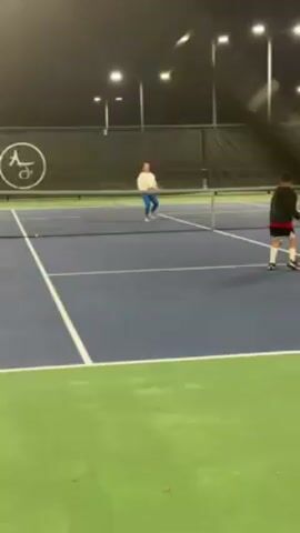 The video shows a woman and a child playing tennis on an outdoor court at night. The court is blue with green borders. The woman, wearing a white shirt and blue pants, is on one side of the net, while the child, wearing a black jacket, red shorts, and white socks, is on the other. The camera is positioned behind a chain-link fence. The child hits a tennis ball over the net. There are several tennis balls on the court. The scene is lit by overhead lights.