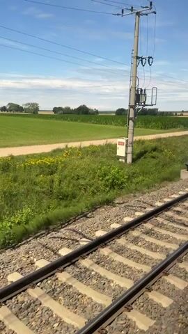 The video shows a view from a moving train. In the foreground, the train tracks and ballast are visible. Beyond the tracks, there are fields of green grass and crops, separated by a dirt path. In the background, more fields stretch to the horizon, dotted with trees. The sky is blue with some scattered clouds. There are utility poles and wires visible along the side of the tracks. The camera appears to be mounted on the side of the train, offering a panoramic view of the passing landscape. The train appears to be moving at a moderate speed, causing a slight blur in the foreground.