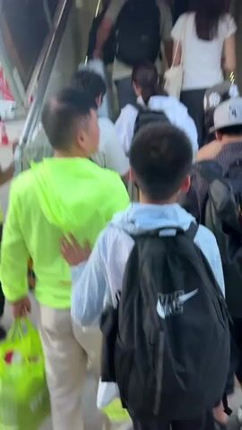 A group of people are walking up a covered staircase or ramp leading to an airplane. The staircase is enclosed with curved metal framing and lighting. The individuals are carrying backpacks and bags. One person in the foreground is wearing a bright green hooded jacket. Another person is wearing a black backpack with a white Nike logo. The lighting is somewhat dim inside the enclosed walkway, with brighter light visible at the top. The ground outside the walkway shows glimpses of pavement, some vehicles, and a red structure that might be part of the airport infrastructure. The people are in single file, ascending the walkway.