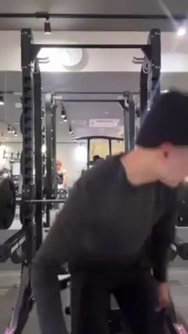 The video shows a person in a gym using a weight bench for a barbell bench press exercise. The person, wearing dark clothes, lies on the bench, grips the barbell, and performs the exercise, lifting and lowering the weight. The bench and barbell are part of a weight rack system with multiple levels. The gym is well-lit, with mirrors, other equipment, and a couple of other people visible in the background.