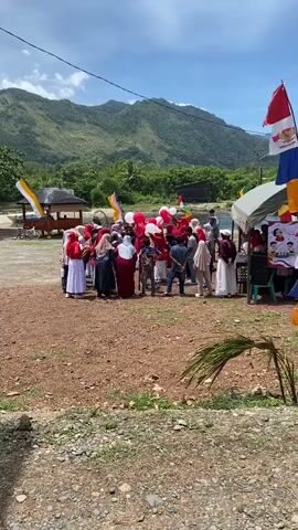 The video captures an outdoor gathering of people, likely a celebration or event, on a sunny day with a backdrop of lush green mountains. A group of individuals, many wearing red and white attire, are clustered together. Some are holding red and white balloons. Flags, including what appears to be the Indonesian flag (red and white), are visible, adding to the festive atmosphere. There's a small structure or stall to the left and a table with a banner to the right, possibly related to the event. The ground is covered in gravel and some dry grass. The overall scene suggests a community event taking place in a natural, somewhat rural setting.
