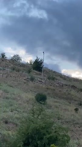 The video displays a static wide shot of a dry, grassy hillside dotted with sparse green bushes and rocks under an overcast sky. The camera slowly pans right, revealing a series of tall, slender streetlights positioned at intervals along the rising terrain. A bird is visible perched atop at least two of these streetlights. The sky is largely covered by dark clouds, with a small patch of brighter sky visible towards the right horizon.