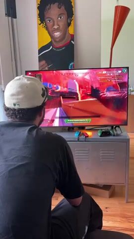A man is seated on a wooden floor, viewed from behind, wearing a dark t-shirt and a light-colored baseball cap with a green logo. He is focused on a large flat-screen television displaying a video game, which appears to be a first-person shooter or battle royale game with French text visible on the interface (e.g., "ÉLIMINATION").

The TV sits on a grey metal cabinet, below which are various game controllers, including red and blue Nintendo Switch Joy-Cons and a green Xbox controller. On the wall above the television, a vibrant painting of a dark-skinned man with curly hair, resembling Ronaldinho, is prominently displayed. To the right of the TV, an orange, conical lamp or sculpture adds a decorative touch. The room has white walls and is well-lit, suggesting it is daytime. The setting appears to be a living room or entertainment area in Villeurbanne, France.