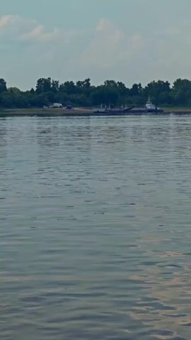 The video begins with a wide shot of a wide river, a tugboat and barge moving in the distance, and trees lining the far bank under a cloudy sky. The camera then pans down and to the right, focusing on the water's surface, showing gentle waves and ripples. As the video progresses, the camera moves closer to the water, revealing the wake behind a moving vessel. The video ends with a close-up shot of the disturbed water and a portion of the vessel's wooden railing in the foreground.