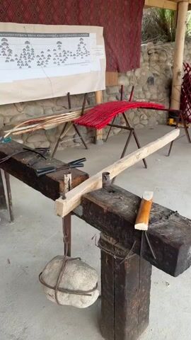The video shows a close-up of a traditional woodworking setup, possibly for crafting bows or similar items. In the foreground, a thick wooden beam rests on two dark wooden supports. This beam has a slot carved into it, and a lighter wooden piece is positioned within this slot, held in place by a metal clamp. Attached to the left side of the main beam is another wooden piece, suspended by a rope connected to a large stone. Various tools, including what appear to be metal blades or clamps, are visible on the main beam. To the right, another wooden support is visible with a carved piece of wood resting on it. In the background, there's a large sheet of paper with diagrams and text, possibly a genealogical chart or a technical drawing. Behind that, a red and white textile hangs on the wall, and a stack of wooden staves is arranged on a metal stand. The overall environment suggests an outdoor or semi-enclosed workshop with a stone wall and a wooden roof structure.
