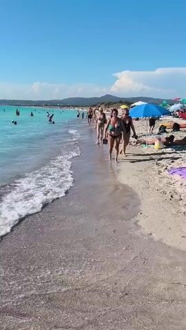 The video shows a beach scene on a sunny day. The water is a clear turquoise color, and there are people swimming and relaxing in it. The beach is sandy, with many umbrellas and beach chairs set up. In the foreground, two women are walking along the shoreline, both wearing bikinis and carrying flip-flops. They are walking towards the right side of the frame. Behind them, other people are visible on the beach, and in the distance, there are hills. A rack of clothing is hanging on the right side of the frame in the foreground.