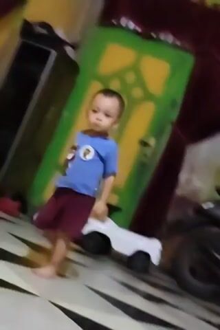 A toddler in a blue t-shirt and maroon shorts walks and dances on a floor with a black and white geometric pattern. The walls are yellow and blue, with a dark red curtain. The toddler is barefoot and appears to be alone.