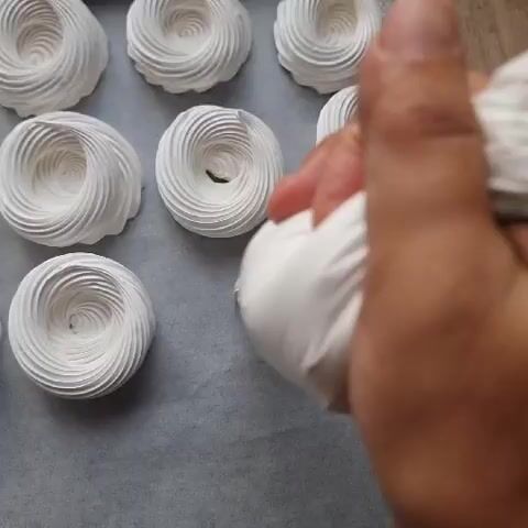A close-up shot shows a person's hand squeezing a piping bag filled with white frosting onto a baking sheet lined with parchment paper. There are several completed meringue nests already on the baking sheet, arranged in a grid. The person is piping a new meringue nest onto the baking sheet, forming a circular pattern with the frosting. The background is blurred, but suggests a wooden surface. The lighting is bright and even.