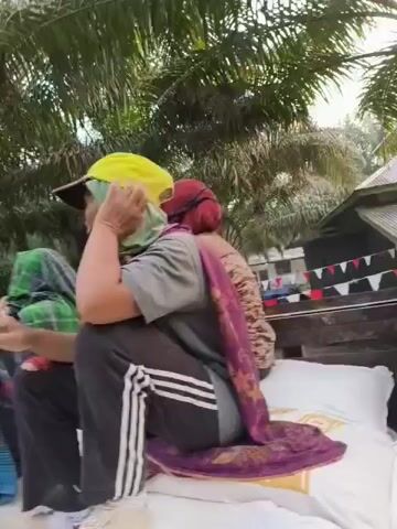 In the video, several people are seen sitting on what appears to be the back of a truck or a similar vehicle, surrounded by palm trees. The individuals are dressed in casual clothing, with some wearing head coverings or face masks. One person is wearing a yellow cap and a grey shirt with black pants. Another is wearing a bright orange vest over a grey and yellow shirt, and another person wears a green and yellow vest. The overall environment suggests an outdoor setting, possibly a rural or agricultural area, with lush greenery and a cloudy sky. The camera is positioned from behind some of the individuals, looking forward. The overall impression is of people being transported or gathered in a natural, possibly working, environment.