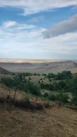 A vertical video displays a scenic landscape. In the foreground, dry, brown grass covers the ground, with a wire fence running horizontally. Beyond this, a valley unfolds with green trees and shrubs scattered across rolling hills. A paved road winds through the valley, leading towards a distant, flat plain. The plain is dotted with small structures, possibly buildings, and power lines stretch across the horizon. The background features a range of soft, undulating hills under a sky filled with white clouds against a pale blue backdrop. The overall impression is one of a rural, open landscape.