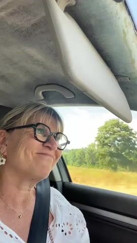 A woman with blonde hair and glasses is sitting in the passenger seat of a car. She is wearing a white shirt with lace trim and a seatbelt. The car is moving, and the scenery outside the window appears to be a rural landscape with trees and fields. She looks at the camera and smiles.
