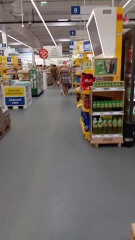 The video captures an interior view of a well-lit supermarket aisle. The floor is a smooth, grey material. Overhead, white fluorescent lights run in a linear pattern, illuminating rows of products on shelves. Signs with blue and yellow accents are visible, indicating aisle numbers and product information. One prominent sign reads "Clubcard Ceny" which translates to "Clubcard Prices". In the distance, several shoppers are present, some pushing shopping carts. On the right side of the aisle, shelves are stocked with numerous green bottles, likely beverages. On the left, a display box with blue and yellow branding is also visible. The overall impression is a typical retail environment.