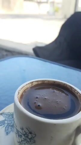 A cup of coffee sits on a blue glass table, its surface reflecting the light. In the background, a black folding chair is positioned next to a gravel area. Beyond the chair, a street can be seen with a white van parked on the side. The scene is outdoors, suggesting a cafe or outdoor seating area.