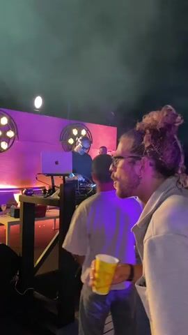 The video shows a man with his hair in a bun, wearing glasses and a grey jacket, holding a yellow cup. He is standing in front of a DJ set where another person is seen working on a laptop. There are lights and smoke in the background, suggesting an event or party. The man appears to be looking to his right. The camera focuses on him, and he is partially obscured by smoke.