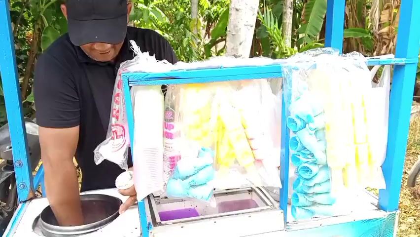 A man wearing a black shirt and a black baseball cap is standing behind a blue and white ice cream cart. He is smiling and looking down at the cart. In front of him, on the cart, is a round, silver metal bowl, which he appears to be interacting with. The cart is filled with various ice cream products in colorful packaging, including pink cups, blue and yellow cone-shaped treats, and pink and yellow popsicle-like items. There are also containers of what appear to be purple ice cream or sorbet in the lower section of the cart. The background is out of focus but suggests an outdoor environment with green foliage.