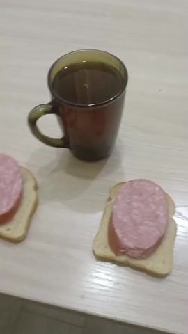 Two sandwiches, each consisting of a slice of white bread topped with a thick slice of pink sausage, are laid out on a light-colored wooden surface. To the left of the sandwiches sits a dark brown, translucent glass mug filled with a dark liquid, presumably tea or coffee. The mug has a visible handle. The scene appears to be indoors, possibly a kitchen or dining area, given the tabletop. The lighting suggests it could be daytime, though the specific time is not clear. No people are visible in the frame. The focus is on the food and drink.
