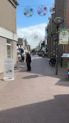 The video shows a sunny daytime scene on a pedestrian street in Noordwijk, Netherlands. The street is paved with reddish bricks in a herringbone pattern, flanked by buildings on both sides. Overhead, decorative, circular ornaments with leaf and fruit motifs are strung across the street.

Initially, an older couple walks into the frame from the left side of the street. The man, wearing a dark jacket, uses a walking cane, and the woman walks beside him. They walk past a white sign that reads "COFFEE AAN ZEE" with a tagline "The only thing better than a cup of coffee is 2 cups of coffee" and illustrations of coffee cups and muffins. As they walk past the camera, other pedestrians can be seen further down the street, some gathered near shops.

On the right side of the street, there's a lamppost with a hanging planter of purple flowers and a banner that says "Welkom, met een glimlach" (Welcome, with a smile) and "Noordwijk aan zee," along with a graphic of a beach scene. Two bicycles are parked against the wall near the lamppost. The sky is bright blue with scattered white clouds, and a small bird can be seen flying overhead at one point. The scene depicts a relaxed atmosphere in a European town center.