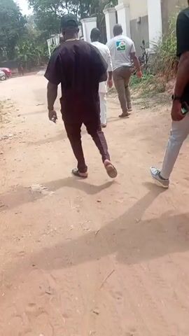 The video shows a group of people walking away from the camera on a dirt path. There are at least six people visible, dressed in various casual outfits. One individual is wearing a dark purple traditional attire and slippers, another a white t-shirt and khaki pants, and another a black t-shirt and light grey jeans. Some are wearing hats. The group is walking towards a building with a white fence. In the background, there is some greenery and a parked motorcycle. The setting appears to be a residential area on a sunny day.