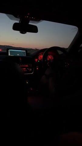 The camera is positioned inside a car, facing forward and slightly to the right, showing the dashboard, steering wheel, and the view through the front windshield. The dashboard lights and instruments glow red, and a digital display on the center console shows a map. A person's hand is visible on the steering wheel on the right side of the frame. Outside the windshield, the sky is a dusky blue and orange, suggesting sunset or sunrise. The landscape appears to be open, possibly a highway, with distant mountains or hills on the horizon. The overall lighting inside the car is dark, highlighting the illuminated dashboard elements.
