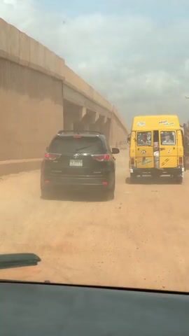 A black SUV with a license plate that appears to read 'FKT 777' is driving down a dirt road. The SUV is kicking up a large amount of dust, obscuring the view of the road ahead. To the right of the SUV is a bright yellow bus with "BRIGHT PATH" written on the back. The bus also has various stickers and drawings on its exterior. The road appears to be under an overpass or bridge. The sky is overcast and hazy, likely due to the dust.