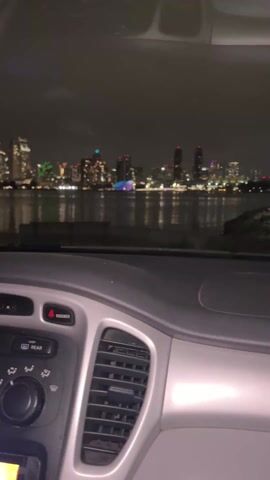 The video shows a view from inside a car, parked, looking out at a brightly lit skyline across a body of water at night. The car's dashboard, with its vents and climate controls, is visible in the foreground. A pink air freshener with the words "LUV is in the air." hangs from the rearview mirror. The time displayed on the car radio is 8:49. The skyline features multiple buildings with various lights, some reflecting on the water. There is some green vegetation on the ground on the near side of the water, to the right.