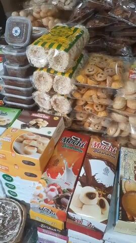 This is a close-up, shaky, handheld video of a market stall piled high with packaged snacks and sweets. The items are predominantly in plastic packaging, with some in cardboard boxes. The snacks appear to be a mix of cookies, wafers, and other dried or fried treats. Several prominent packages of "Mochi" snacks are visible, some with images of milk and chocolate. The stall is crowded with merchandise, creating a dense visual display of food items. The lighting is somewhat dim, with bright highlights reflecting off the plastic packaging. There are no people clearly visible in the frame, and no actions or interactions are captured.