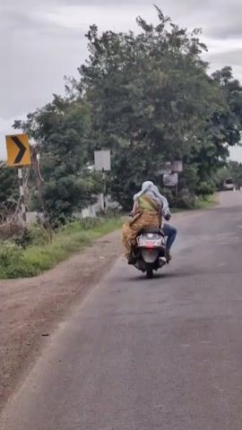 A man and a woman are riding on a scooter. The woman is sitting behind the man and is wearing a sari. They are on a road with trees on either side. The sky is overcast.