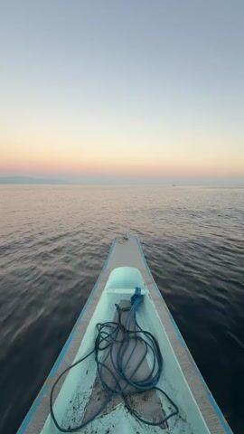 This is a first-person perspective video taken from the bow of a narrow, light blue and grey boat as it glides through calm, dark water. The boat is empty of people. A coiled blue rope lies in the bottom of the boat, secured by a light blue cleat. The horizon is visible in the distance, where the sea meets a soft gradient of pastel colors in the sky, suggesting either sunrise or sunset. The sky transitions from a pale, clear blue at the top to shades of pink, peach, and a faint lavender closer to the horizon. There is a very faint, dark line on the horizon that could be a distant landmass or a very thin cloud formation. The overall mood is peaceful and serene. The video appears to be filmed at dawn or dusk, indicated by the soft, diffused light and the gentle colors in the sky. The setting is a large body of water, likely the sea given the vast horizon. The location context of Buleleng, Indonesia, suggests this could be the Bali Sea or a similar coastal area. There is no visible text in the media file.