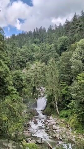 A handheld video pans across a lush, forested mountainside with a rocky river flowing through the center. The river cascades over rocks, creating small waterfalls. Dense green trees and foliage cover the slopes, with a clear sky visible in the upper portion of the frame. The camera moves from left to right, capturing the natural beauty of the landscape. There are no people or man-made objects visible other than the implied presence of the camera operator. The video is likely taken during daylight hours.