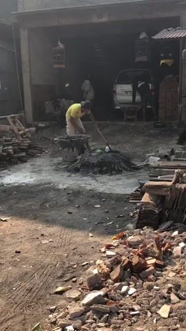 The video shows a construction site with several people working. In the foreground, there is a pile of rubble and debris. In the middle ground, a person in a yellow shirt is using a shovel to mix cement in a wheelbarrow. Another person is visible in the background, near a van. The environment appears to be an open-air workshop or construction area, with materials like bricks and wood stacked around. The overall atmosphere suggests a busy and active worksite.