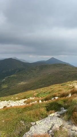 The video shows a panoramic view of mountain ranges under a cloudy sky. The foreground features a grassy slope with scattered rocks, transitioning into lush green slopes and dark forests on the distant mountains. The sky is overcast with dramatic clouds, suggesting a changeable weather. The video slowly pans across the landscape, highlighting the vastness and ruggedness of the mountains.