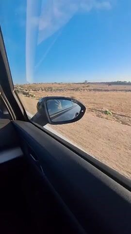 The media file captures a view from inside a car, looking out the passenger side window and through the side mirror. The scene depicts a rural landscape under a clear, bright blue sky, suggesting a sunny day. The ground outside appears to be a dirt road or path, with dry, scrubby vegetation and rocky formations lining its edges. In the distance, more of this arid terrain stretches out, with a few sparse trees or bushes visible. The side mirror reflects a glimpse of the interior of the car, including what appears to be a hand resting on the steering wheel, implying someone is driving. The sky has a few wispy clouds. The overall impression is one of a journey through a dry, open landscape on a pleasant day. There is no visible text in the media.