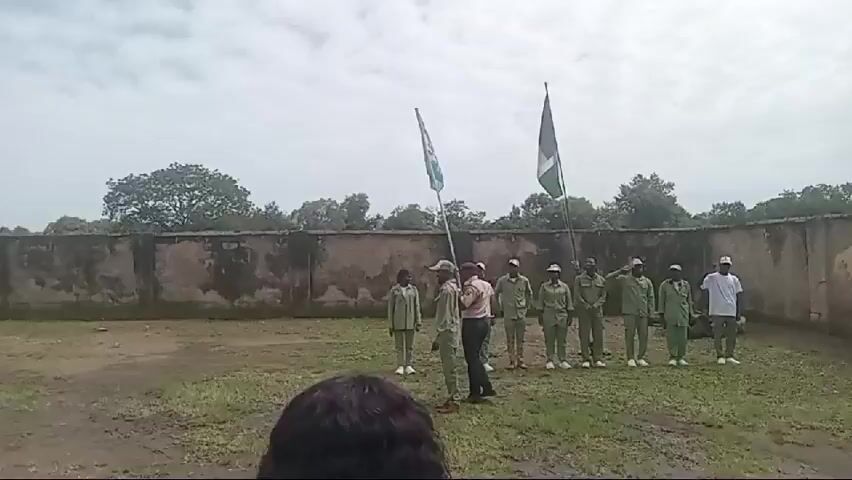 A group of approximately ten individuals, some in green uniforms and others in white shirts and green pants, stand in formation on a grassy field. Two flags are planted on poles in front of them. A person in a light-colored shirt and dark pants stands with their back to the camera, facing the uniformed individuals. Another person, also in a green uniform, holds one of the flags. Several individuals in the formation are saluting. A wall runs along the back of the scene, with trees and foliage visible beyond it. The sky is overcast. The camera appears to be positioned behind another person, whose dark hair is visible in the foreground.