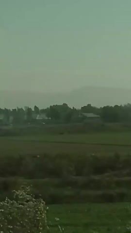 The video shows a landscape on a slightly overcast day. In the foreground there's green foliage, then a field, followed by trees and some small buildings in the distance. Further in the background are faint mountains obscured by haze. The camera remains still during the short video.