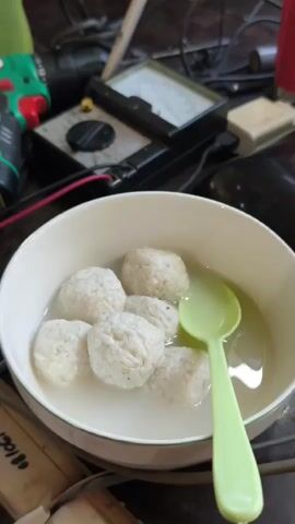 A close-up, slightly blurry, overhead shot shows a white bowl filled with six white, round rice balls submerged in a clear liquid. A light green spoon is partially submerged in the liquid, resting on top of the rice balls. In the background, out of focus, is a black object with red and black wires, and a vintage-looking electrical meter with a dial and knobs. The lighting is somewhat dim.