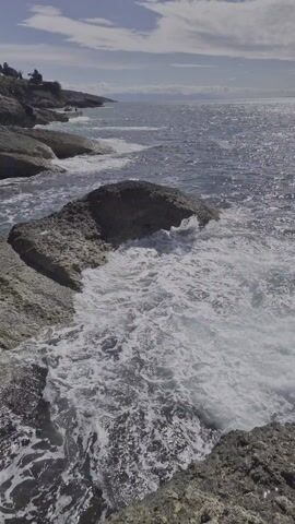 The video shows a rocky coastline with waves crashing against the rocks. The camera pans up to reveal a large stone building, possibly a fortress or castle, perched on top of the rocks. The sky is blue with some clouds.
