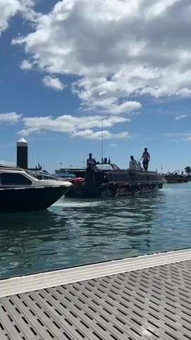 A group of people are on a large, black boat with red accents. The boat has a tan and black cabin structure with windows. One person stands at the stern with their arms raised. Another person stands on the roof of the cabin, and at least two other individuals are visible on the deck of the boat. The boat is moving through clear blue water, leaving a wake. In the foreground, a grey and white dock with metal grating is visible. To the left of the frame, a smaller black and white boat is partially visible. The sky is blue with scattered white clouds. In the background, other boats and structures are visible, suggesting a marina or harbor.