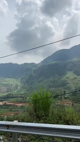 The video captures a wide, sweeping view of a mountainous landscape in Minh Sơn Commune, Vietnam, filmed from a moving vehicle. Lush green mountains dominate the background, covered in dense trees and foliage. In the mid-ground and valleys, terraced fields, possibly agricultural, are visible, along with scattered buildings or a small village.

In the foreground, a metal guardrail runs horizontally, indicating the presence of a road. Green corn stalks and other wild vegetation grow along the roadside, obscuring the view slightly at times. Power lines stretch across the frame, supported by a utility pole visible briefly on the left.

The sky is overcast with large, grey clouds, but the overall lighting is bright, suggesting daytime. The camera pans slowly from left to right, revealing more of the expansive valley and mountain slopes. The predominant sound is wind, indicating the recording device is exposed, likely from a moving vehicle. No people or visible text are present.