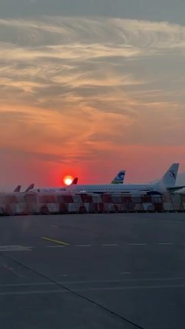 The video shows a sunset behind airplanes parked on a tarmac. The sky is filled with orange and red hues as the sun dips below the horizon, creating a vibrant backdrop for the planes. There are three airplanes visible, parked behind a striped barrier. The runway in front of the planes is grey, marked with yellow lines. In the background, a silhouette of a mountain range can be seen. The camera slowly pans to the right during the duration of the video.
